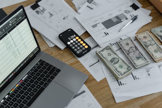 an open laptop next to scattered papers, a calculator, and stacks of cash